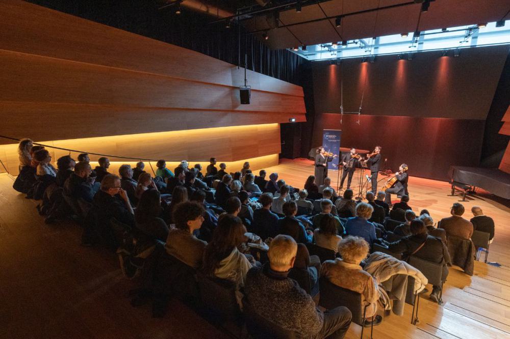 Publikum sitzt in einem modern gestalteten Konzertsaal mit Holzverkleidung und blickt auf ein Streicherquartett, das auf der Bühne spielt.